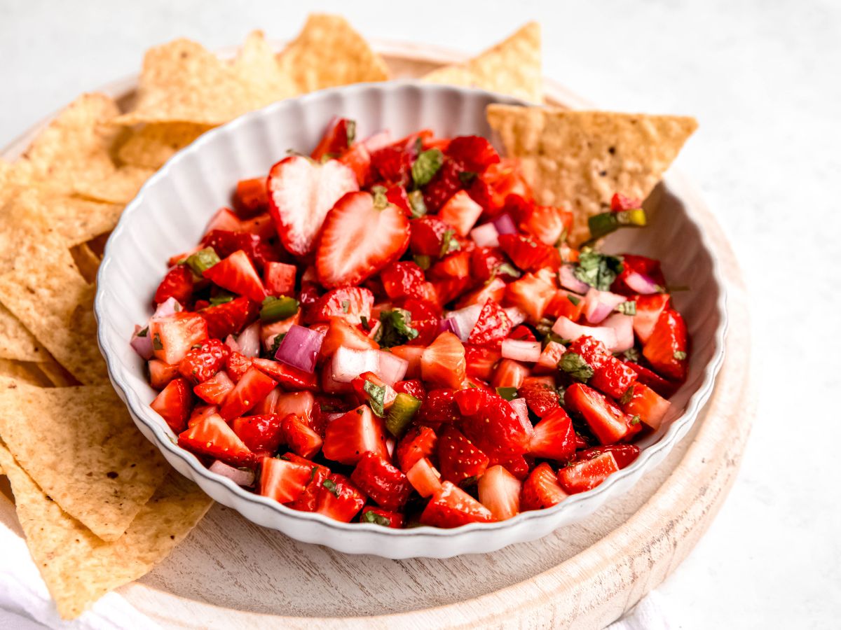 horizontally framed hero shot of strawberry salsa in a bowl on a white wooden serving tray filled with tortilla chips.