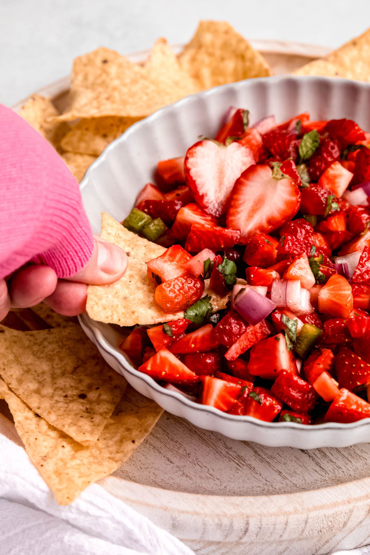 45 degree angle of a hand in a pink sweater up to the knuckles scooping a bite of fresh strawberry salsa with a tortilla chip.