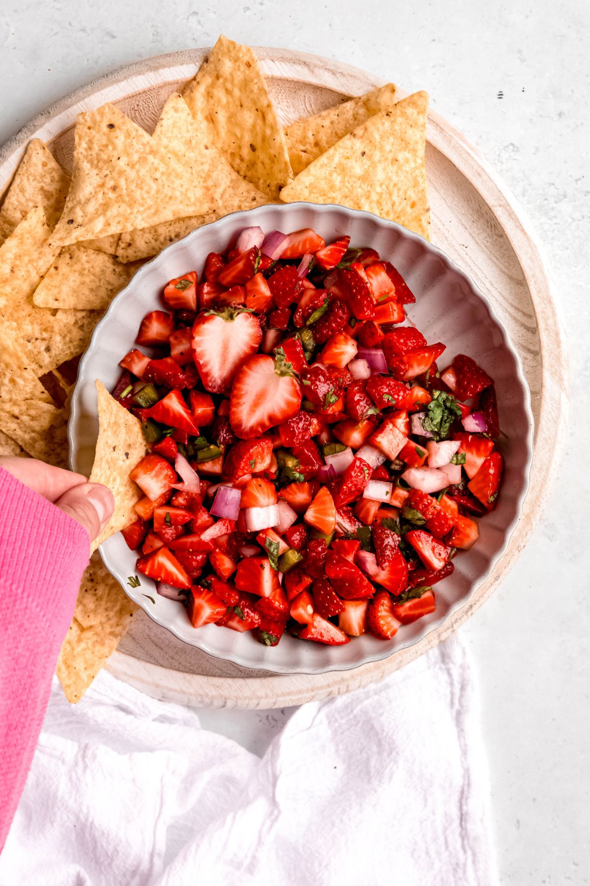 overhead shot of a hand in a pink sweater up to the knuckles dipping a tortilla chip into a bowl of fresh strawberry salsa.