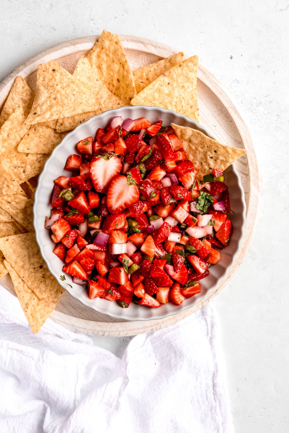 white scalloped bowl filled with strawberry pico de gallo salsa on a white wooden serving tray with tortilla chips on a white table.