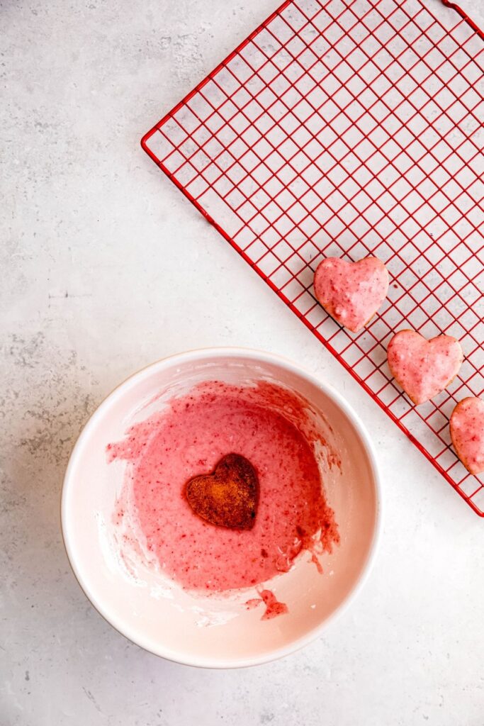 cooled strawberry shortbread hearts being dipped in the strawberry glaze and placed on a wire rack to drip off the excess.