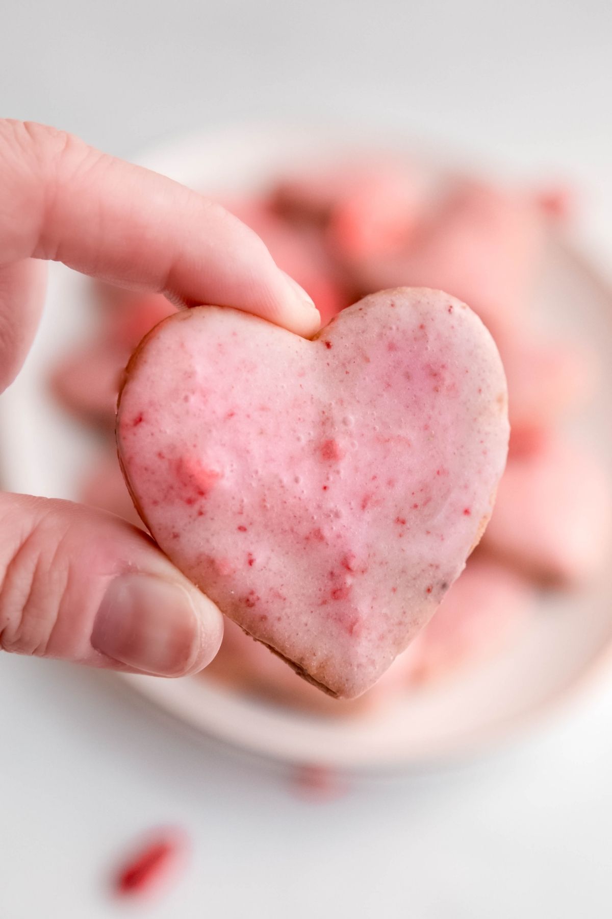 thumb and forefinger holding a single strawberry shortbread cookie to show the pretty pale pink glaze with tiny flecks of strawberries in it; the plate with the rest of the cookies is blurred out in the background.