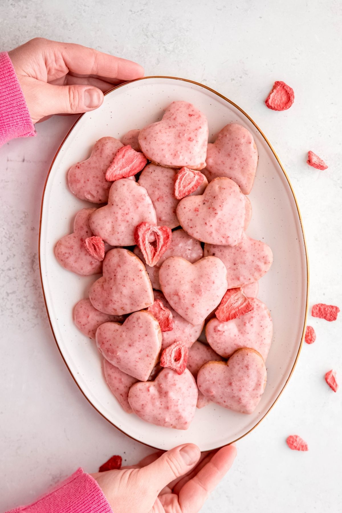hands in a pink sweater up to the knuckles setting down a white oval serving platter filled with homemade strawberry shortbread cookies with strawberry glaze on a white table with freeze dried strawberries scattered around.