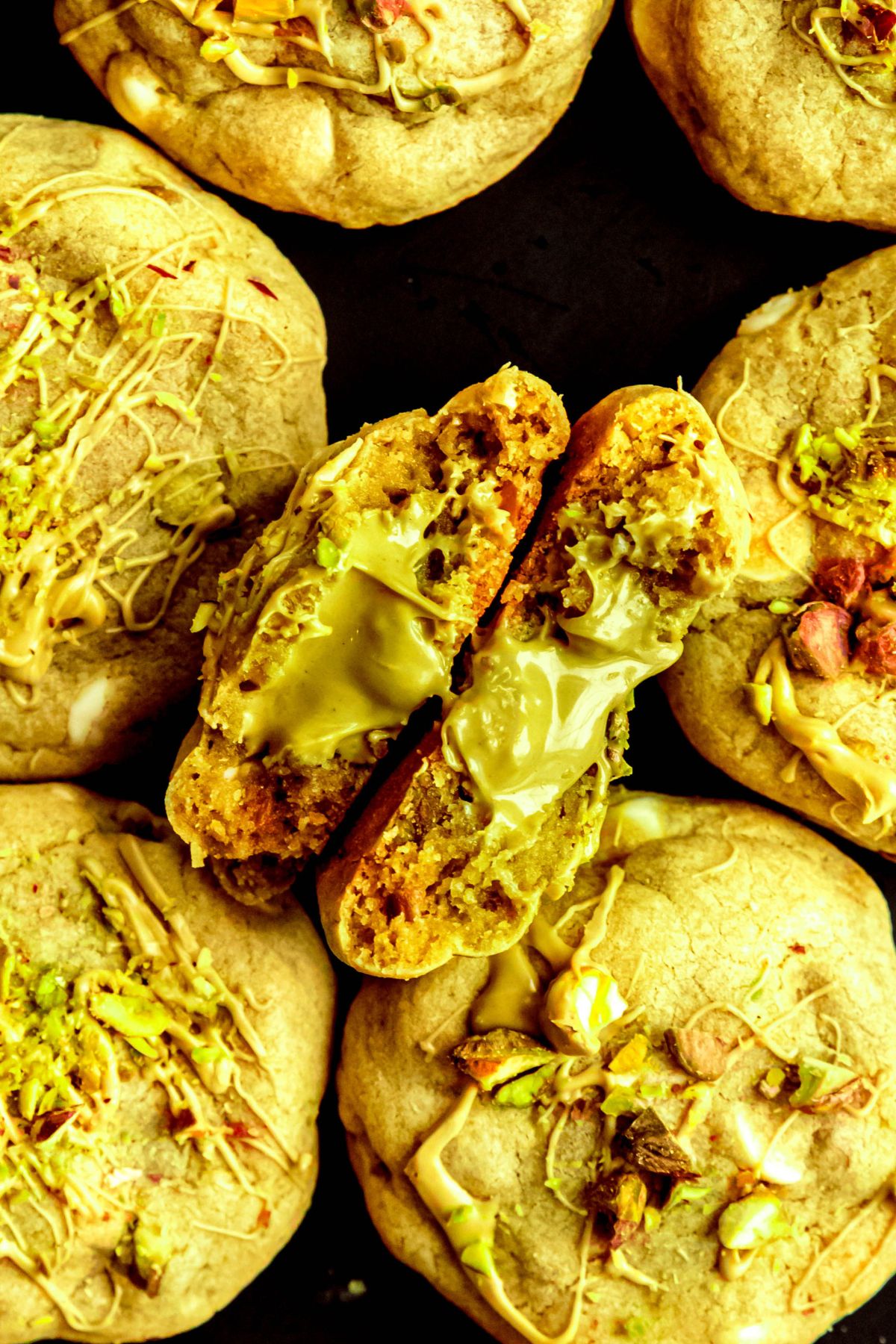 overhead shot of a black plate filled with pistachio cookies with the one in the middle broken in half and facing upwards to show the creamy, melty pocket of pistachio cream filling.