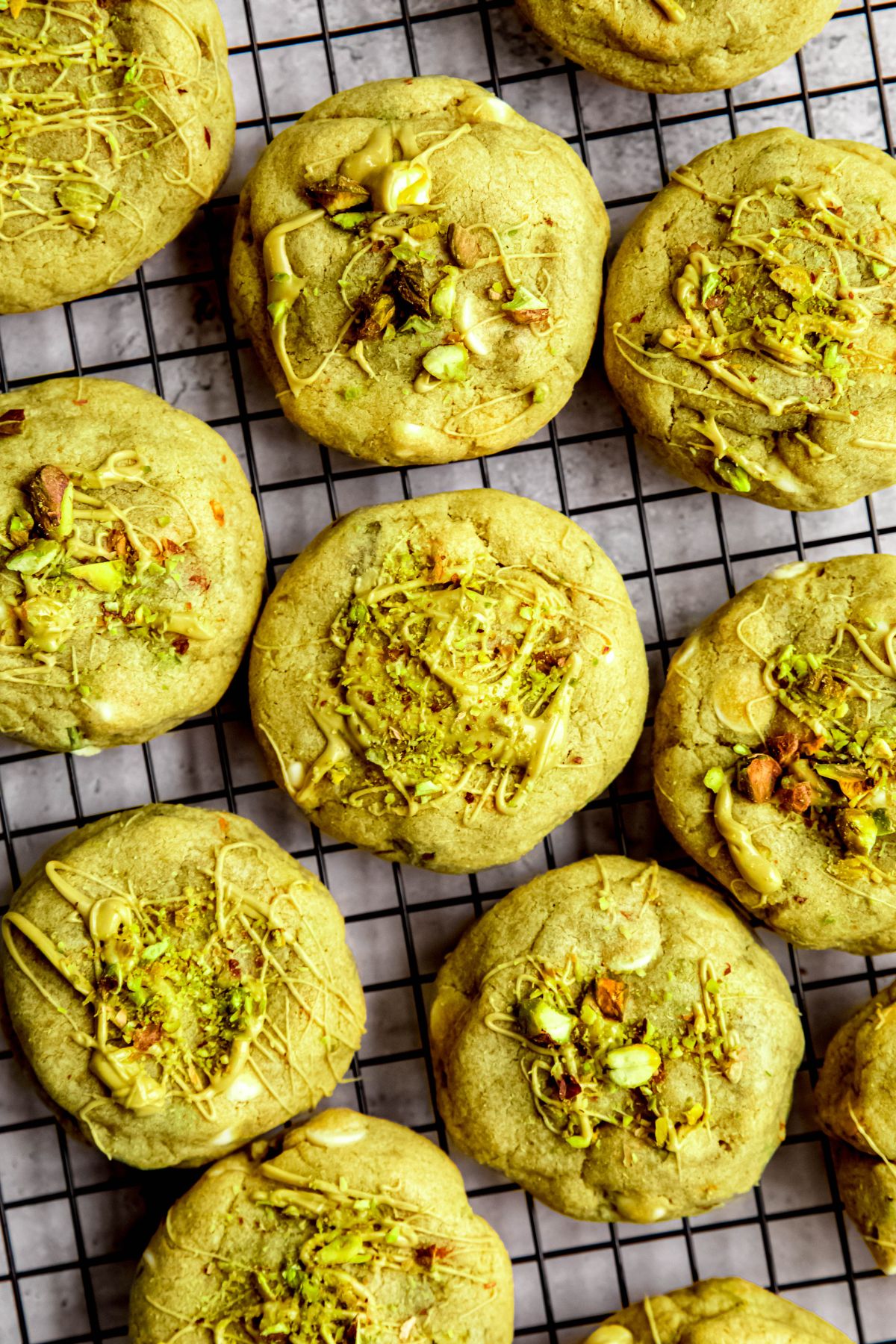 closeup overhead shot of garnished stuffed pistachio cookies on a black cooling rack topped with a drizzle fo pistachio cream and chopped nuts.