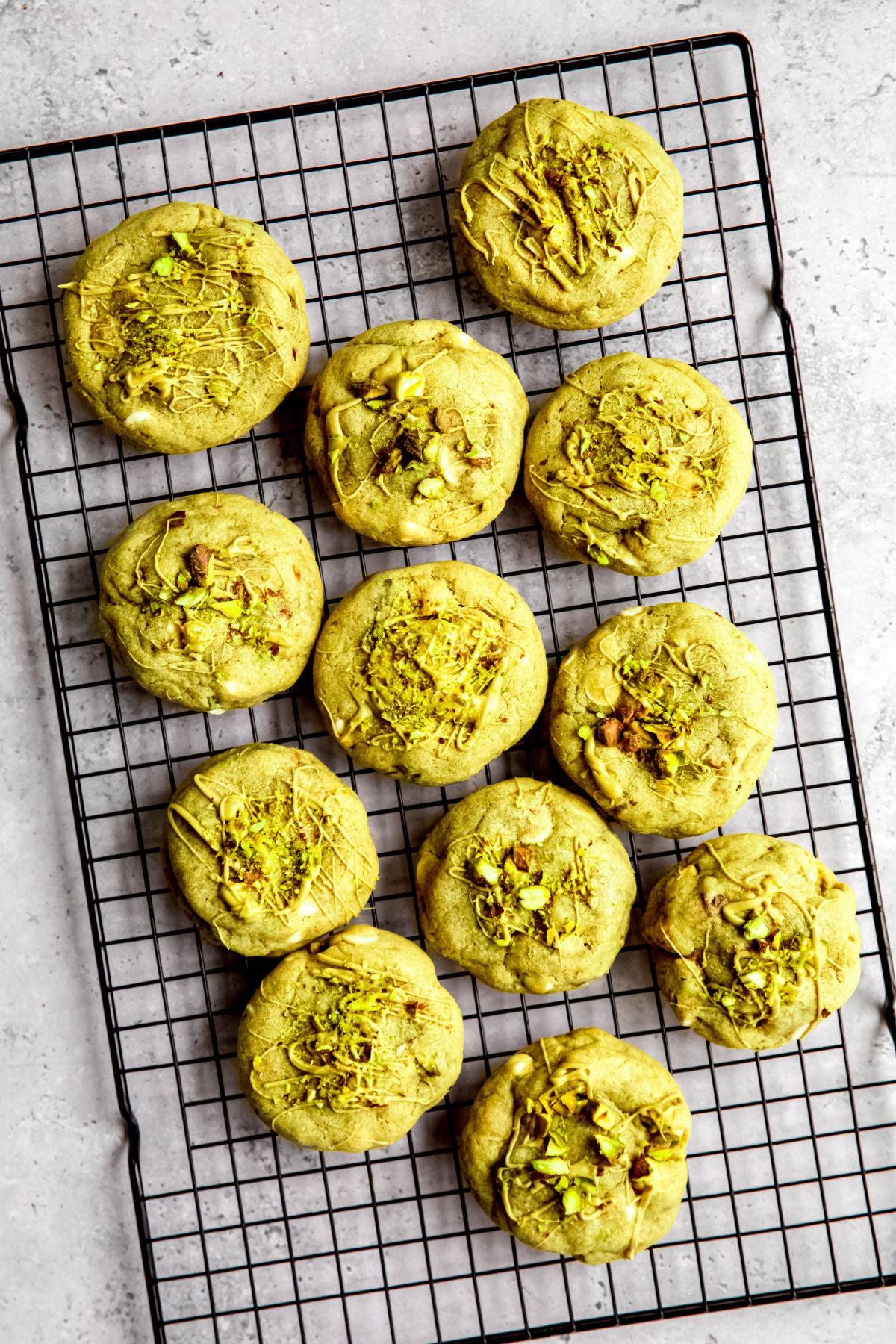 overhead shot of a batch of pistachio cream filled pistachio cookies on a black cooling rack on a grey table.