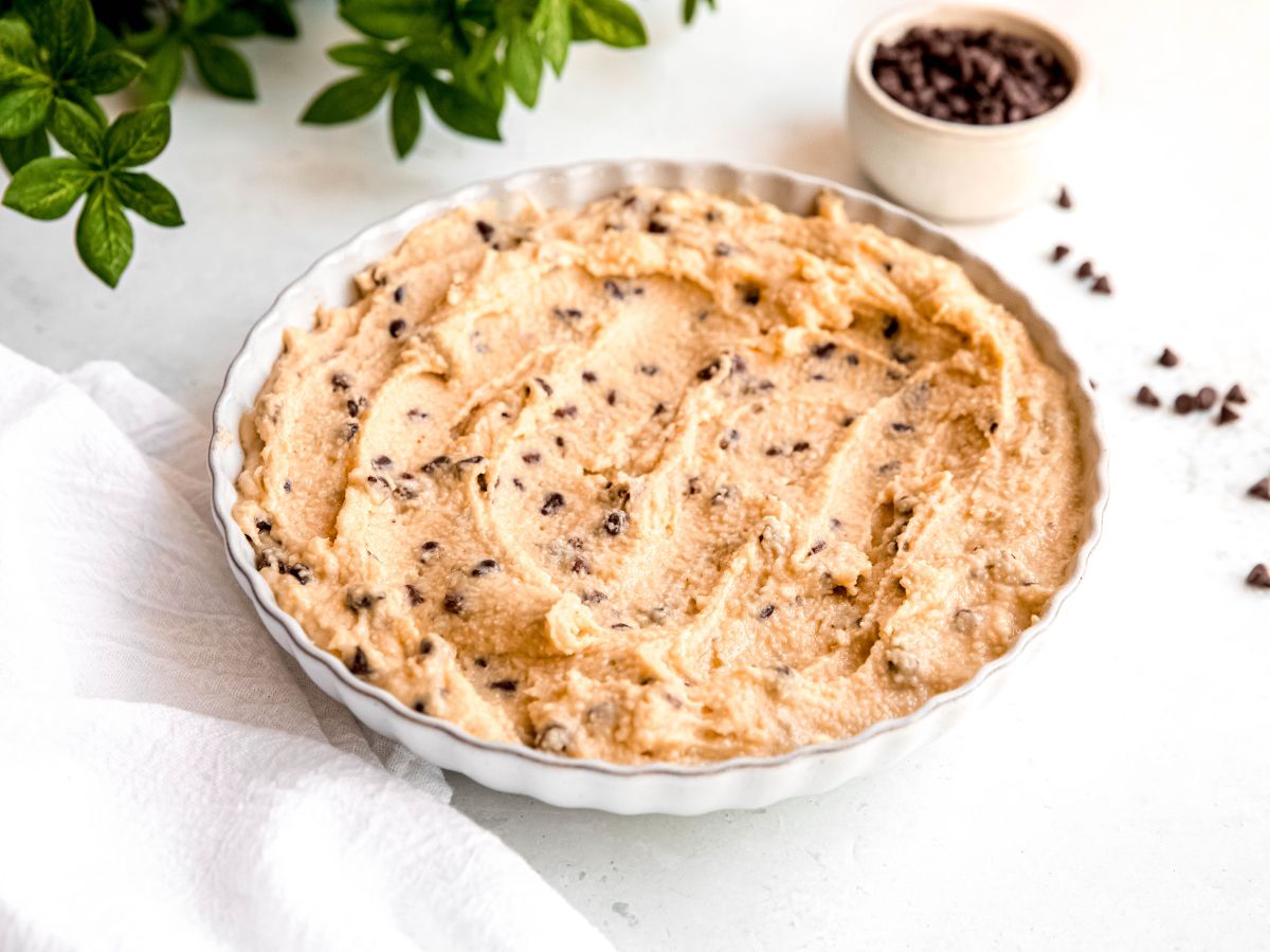 horizontally framed 45 degree angle shot of a white scalloped serving bowl filled with edible cottage cheese protein cookie dough on a white table with a pinch bowl of mini chocolate chips (some of which are scattered around) and a green houseplant.