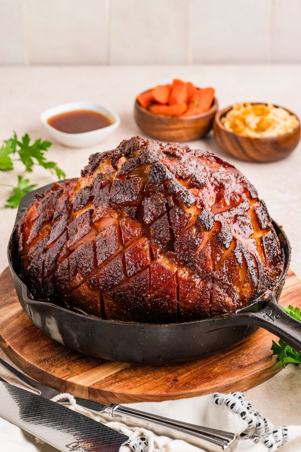 glazed double smoked ham in a cast iron skillet on a wooden trivet on a white table with sprigs of parsley around.