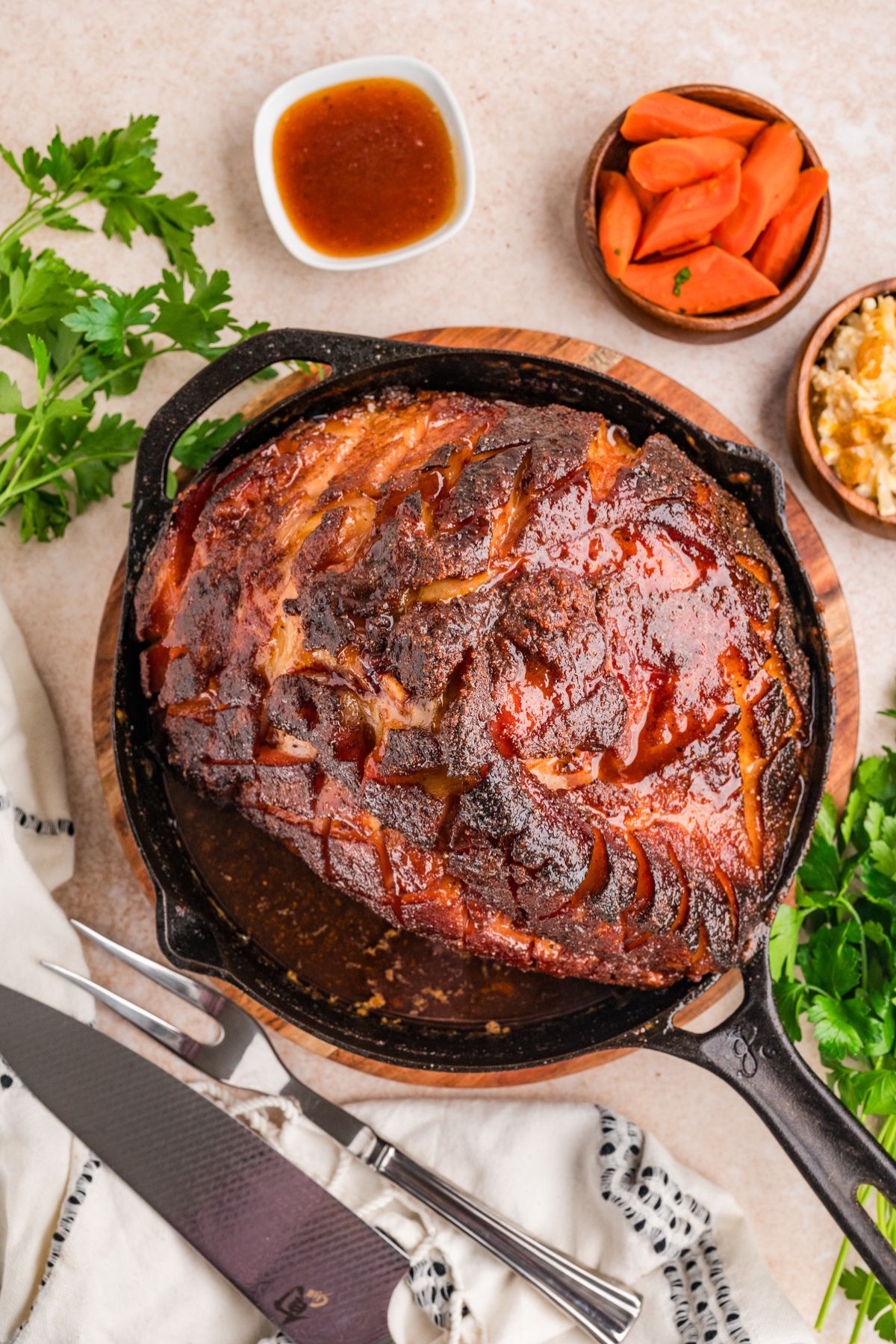 flat lay shot of double smoked ham in a cast iron pan on a wooden trivet on a table with a bunch of parsley and a bowl of honey roasted carrots with a meat fork and chef's knife to the side.