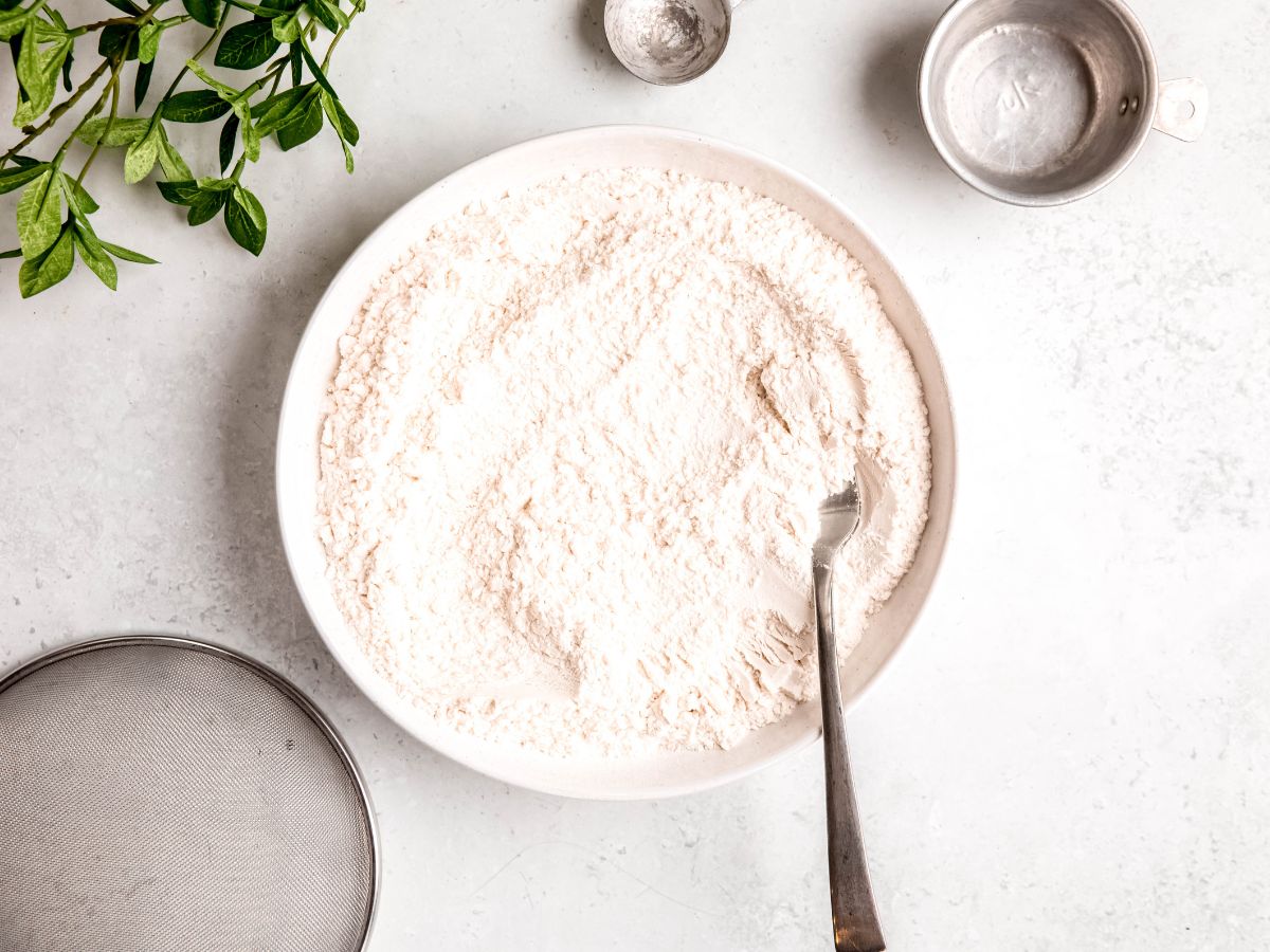 horizontally framed hero shot of self-rising flour made with all-purpose flour on a white table with silver measuring implements and a green houseplant.