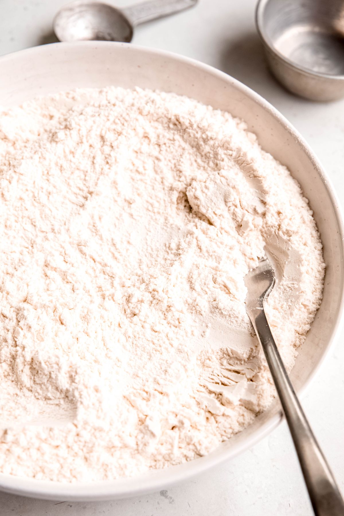 closeup shot of homemade self-rising flour in a white bowl with a silver fork whisking it.