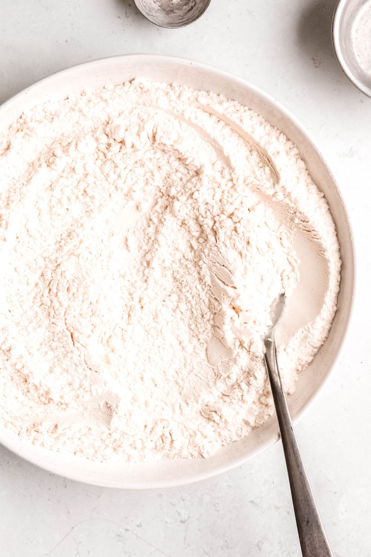 overhead closeup shot of the right 4/5 of a white mixing bowl filled with homemade self rising flour with a silver spoon in it.