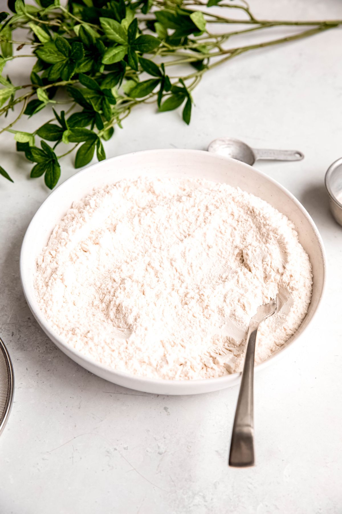 white earthenware bowl filled with DIY self-rising flour on a white table with a mesh sifter, a silver tablespoon measure and a green houseplant.