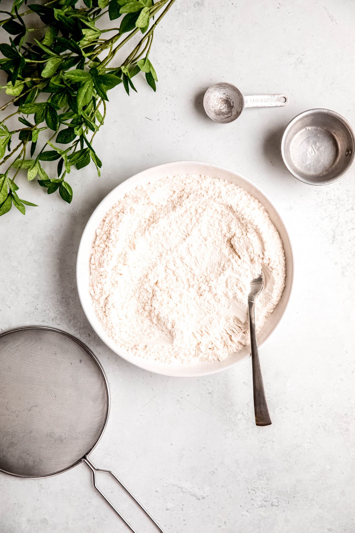 bowl of homemade self rising flour with a silver fork in it on a white table with measuring implements, a mesh sifter, and a green houseplant.