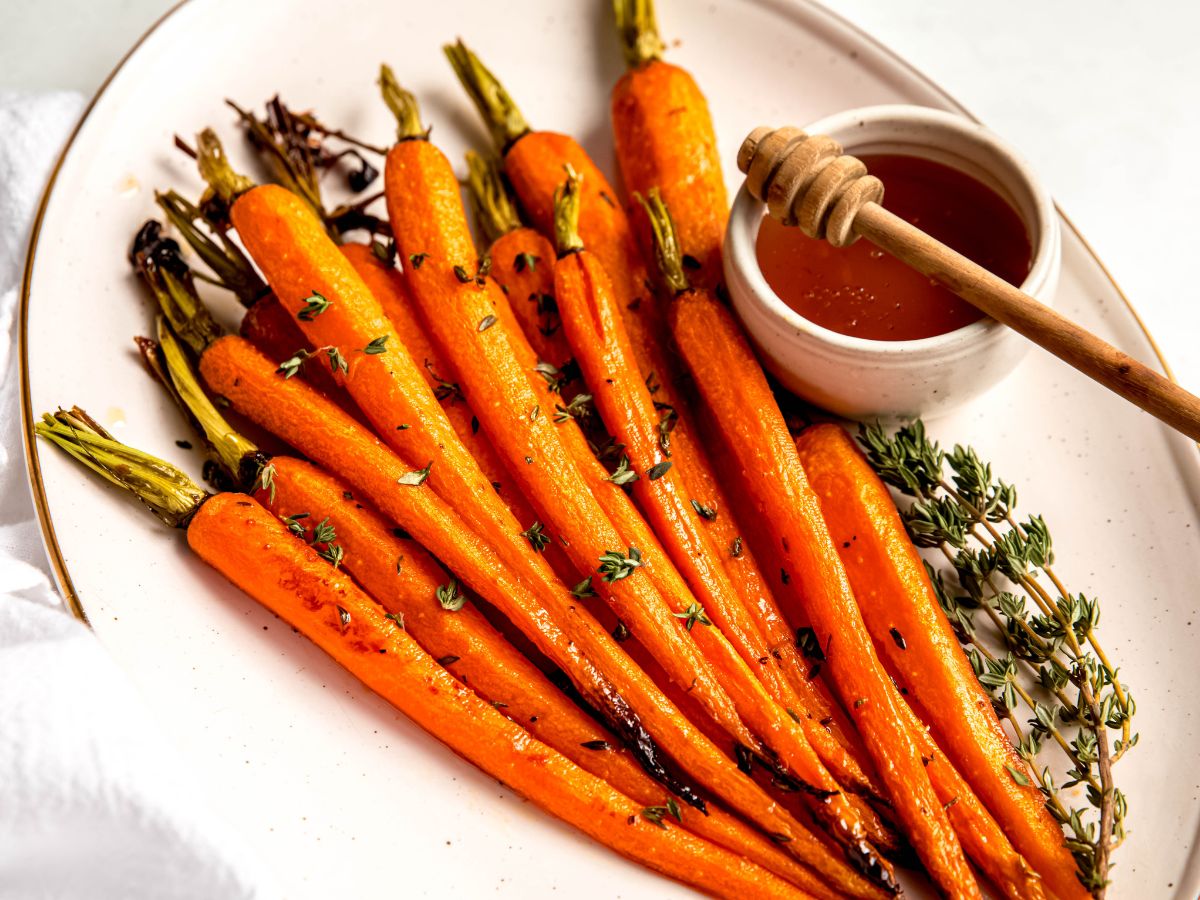 horizontally framed shot of roasted honey and brown sugar glazed carrots on a white oval serving platter with extra fresh thyme and a ramekin of extra glaze with a wooden honey dipper for drizzling.
