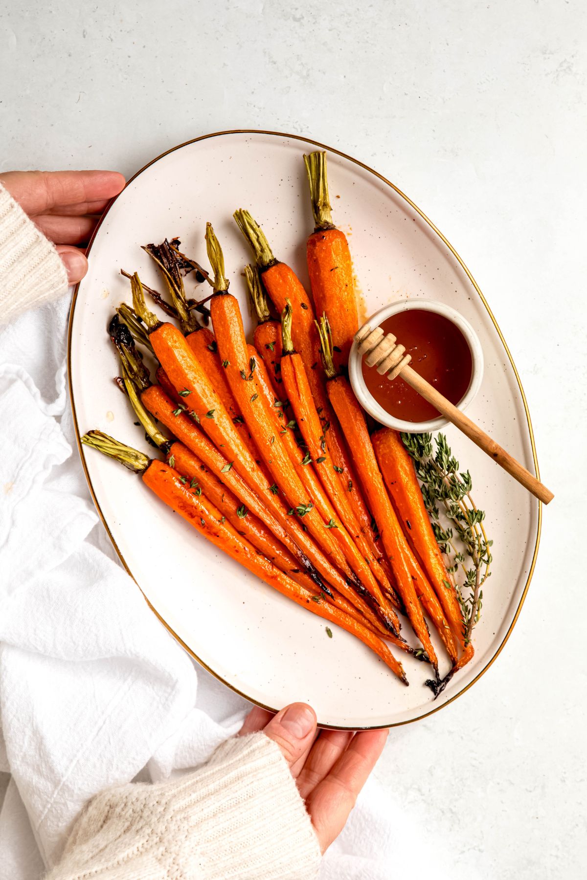 hands in a white sweater up to the knuckles setting down an oval serving platter of honey brown sugar glazed carrots on a white table with a white linen.