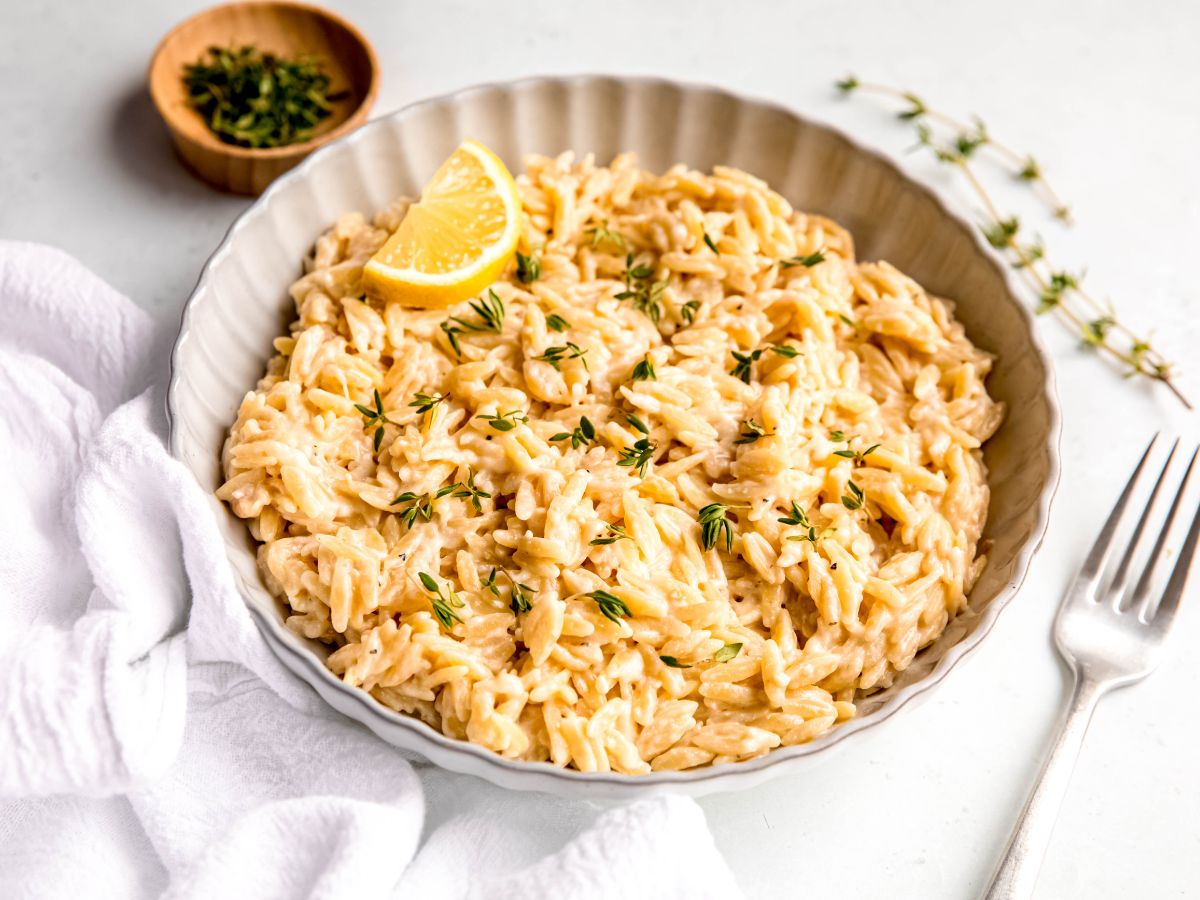horizontally framed hero shot of a white scalloped serving bowl filled with lemon orzo parmesan pasta topped with fresh thyme leaves and a slice of lemon on a white table with a silver fork and fresh sprigs of thyme.