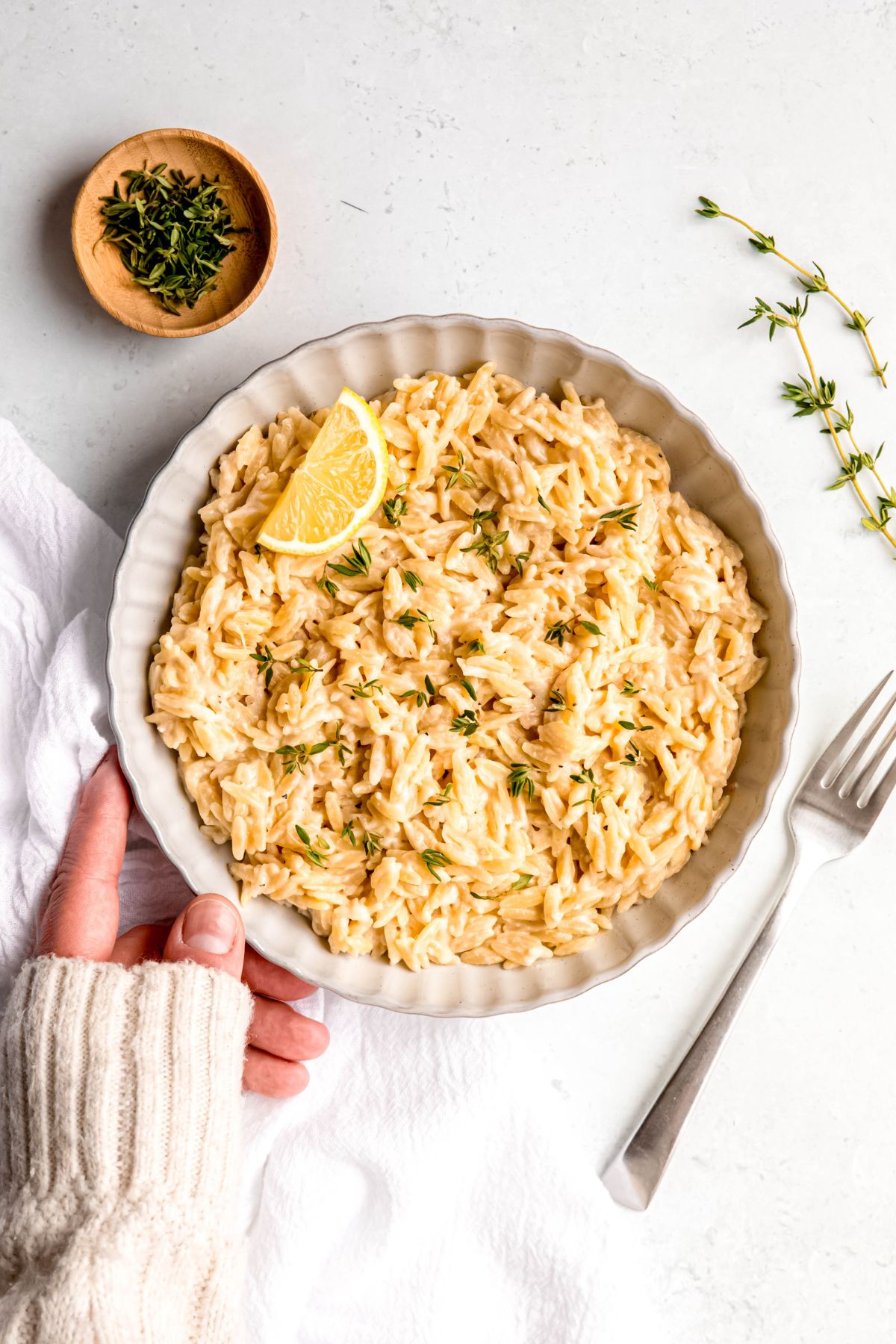 hand in a white sweater up to the knuckles setting down a white serving bowl of lemon orzo pasta on a white table with a silver fork and fresh sprigs of thyme.