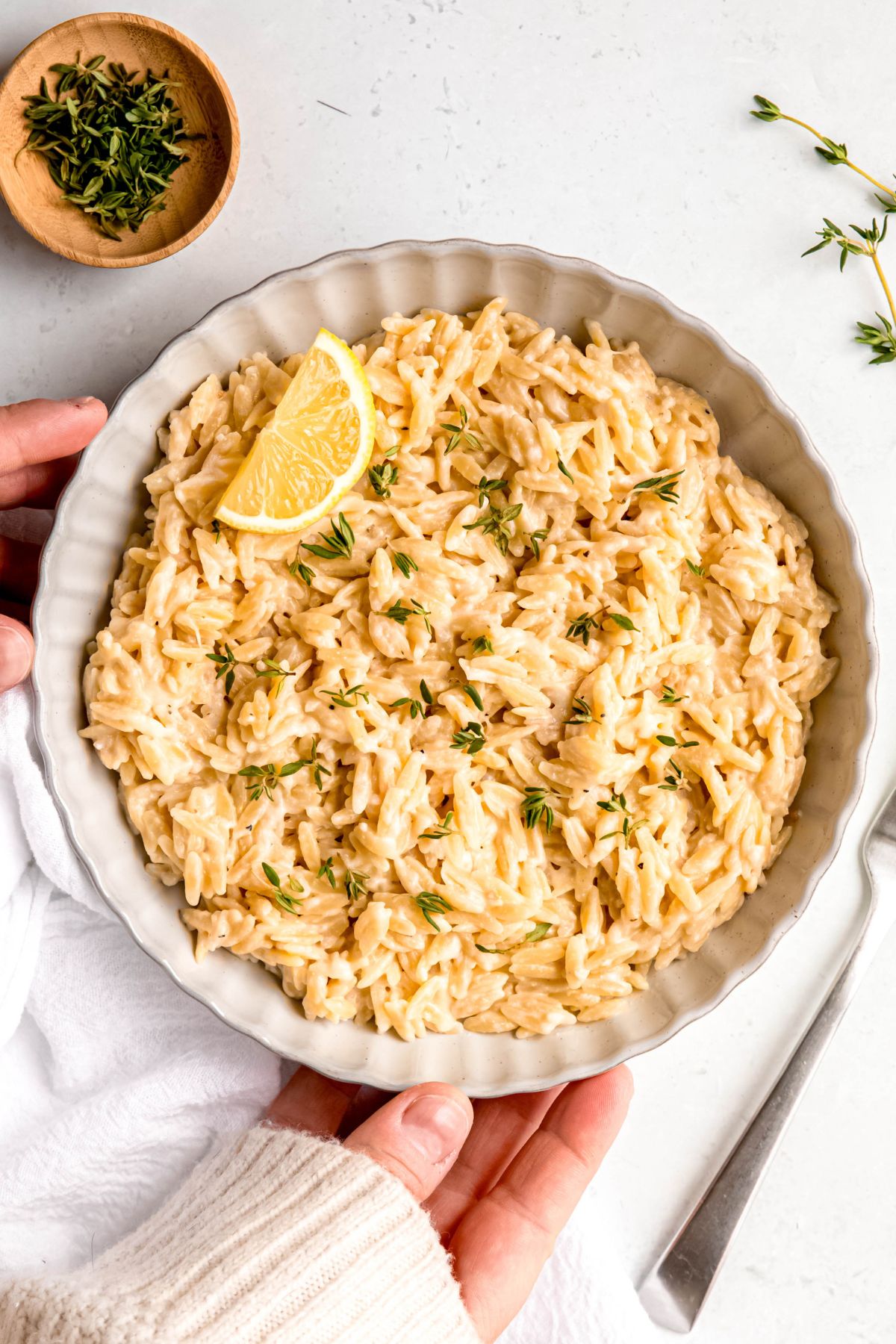 hands setting down a scalloped white serving bowl filled with lemon orzo pasta garnished with a fresh lemon slice and fresh thyme.