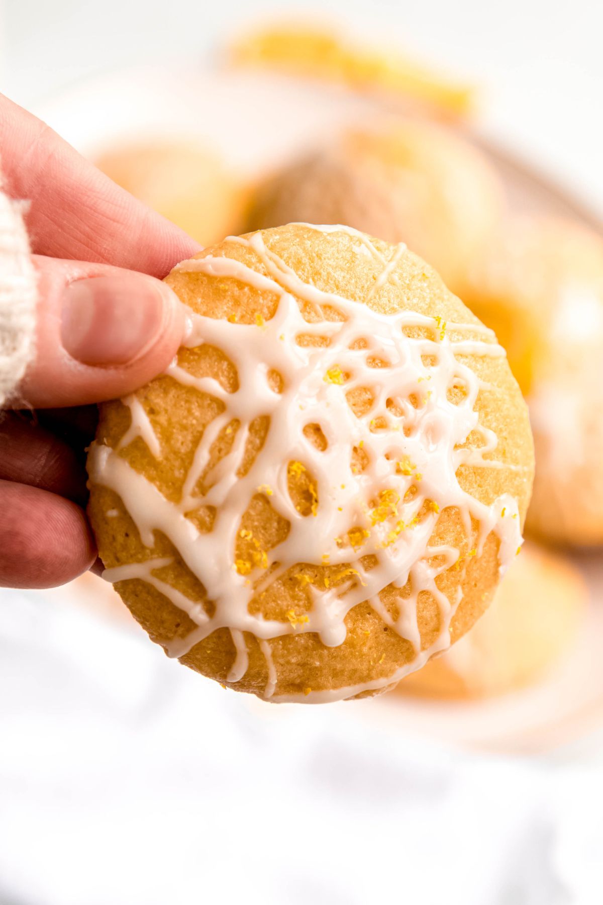 closeup of a hand holding a lemony ricotta cookie with the top facing towards the camera showing the pretty drizzle of white lemon glaze and yellow lemon zest on top.
