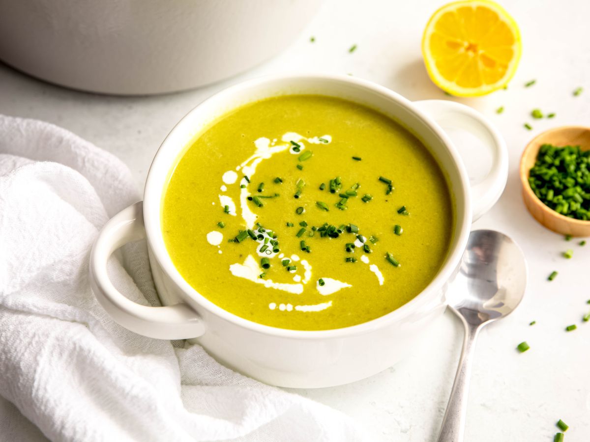 horizontally framed hero shot of a bowl of creamy asparagus soup topped with fresh chives and a drizzle of cream on a white table with a wooden pinch bowl of chopped chives, a halved lemon and a silver spoon.