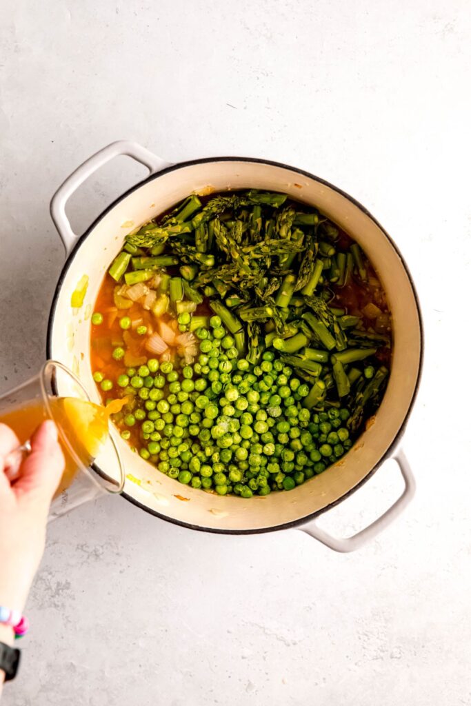 action shot of hand pouring in vegetable broth to the dutch oven after adding the remaining veggies to the sauteed aromatics.
