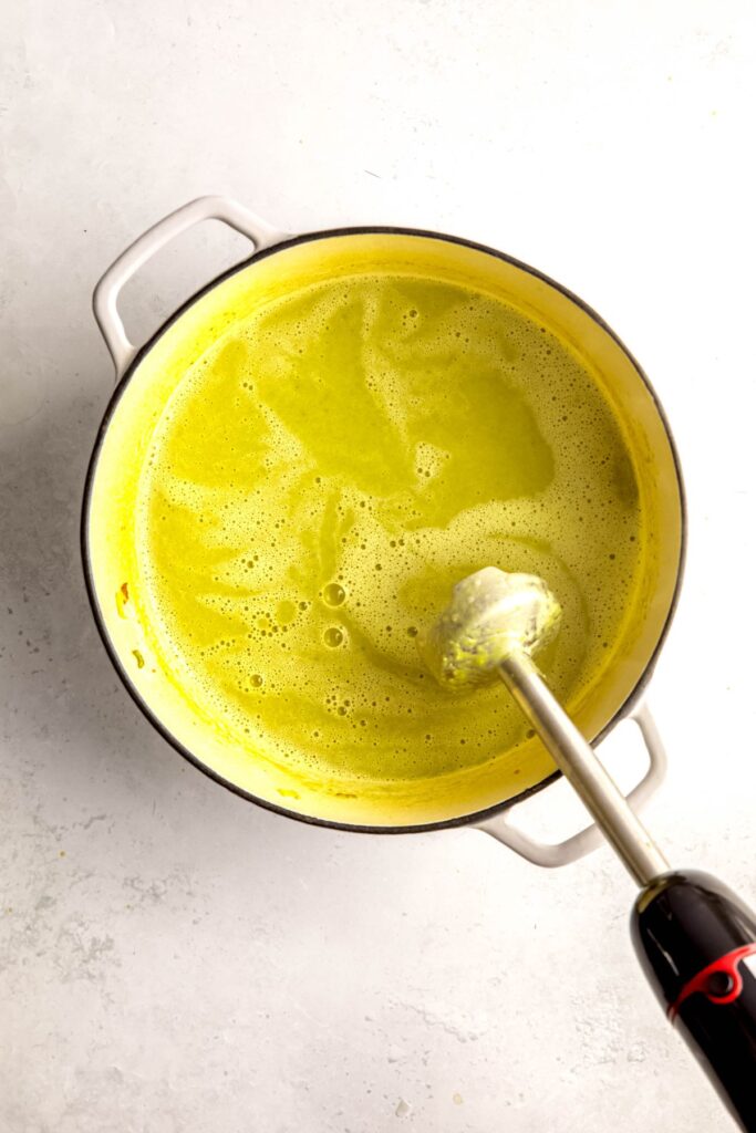 action shot of an immersion blender blending the cream of asparagus soup in the dutch oven until creamy and smooth.