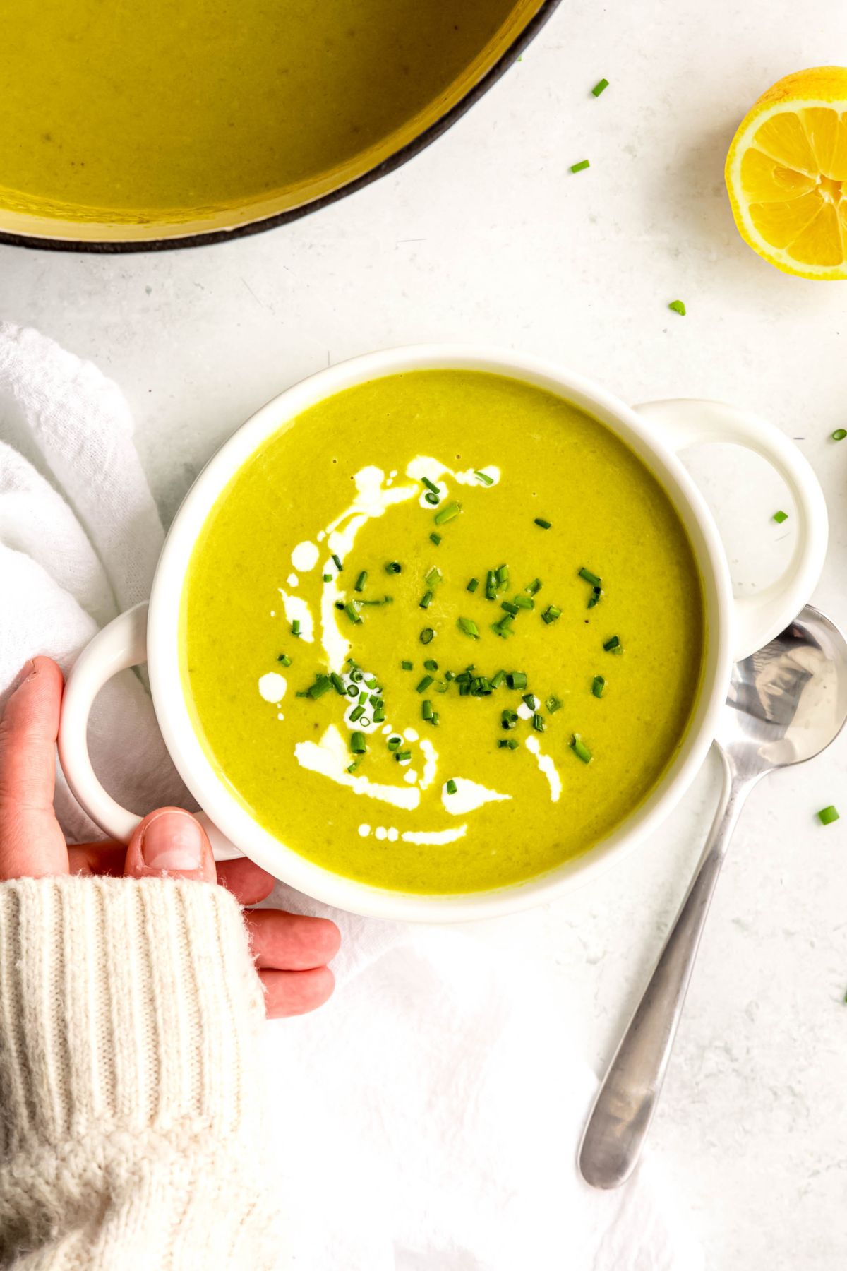 overhead shot of a hand in a white sweater up to the knuckles setting down a white handled bowl filled with cream of asparagus soup on a white table with a silver spoon and a white linen napkin.