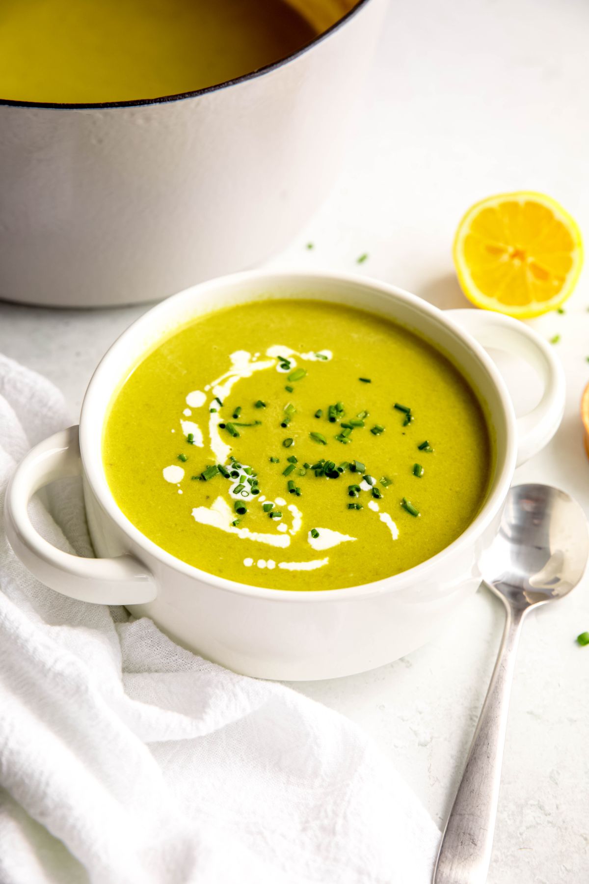 45 degree angle of a white soup bowl with handles filled with a serving of creamed asparagus soup topped with fresh chives and a drizzle of cream on a white table with a silver soup spoon, sliced lemons and a white dutch oven with the rest of the batch of soup.