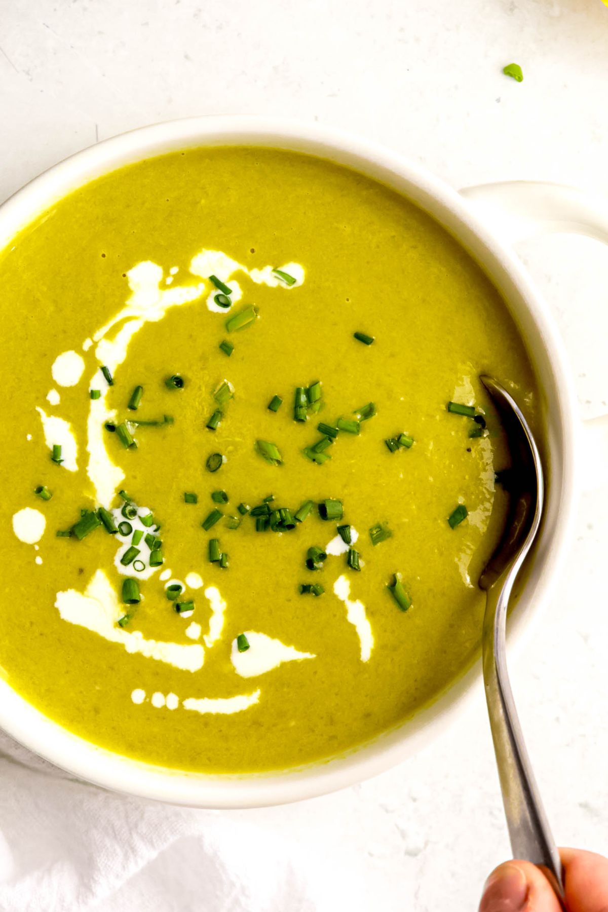 closeup overhead shot of a white bowl filled with cream of asparagus soup on a white table; the bowl is garnished with a drizzle of cream and fresh chives and a hand is digging in with a silver spoon.