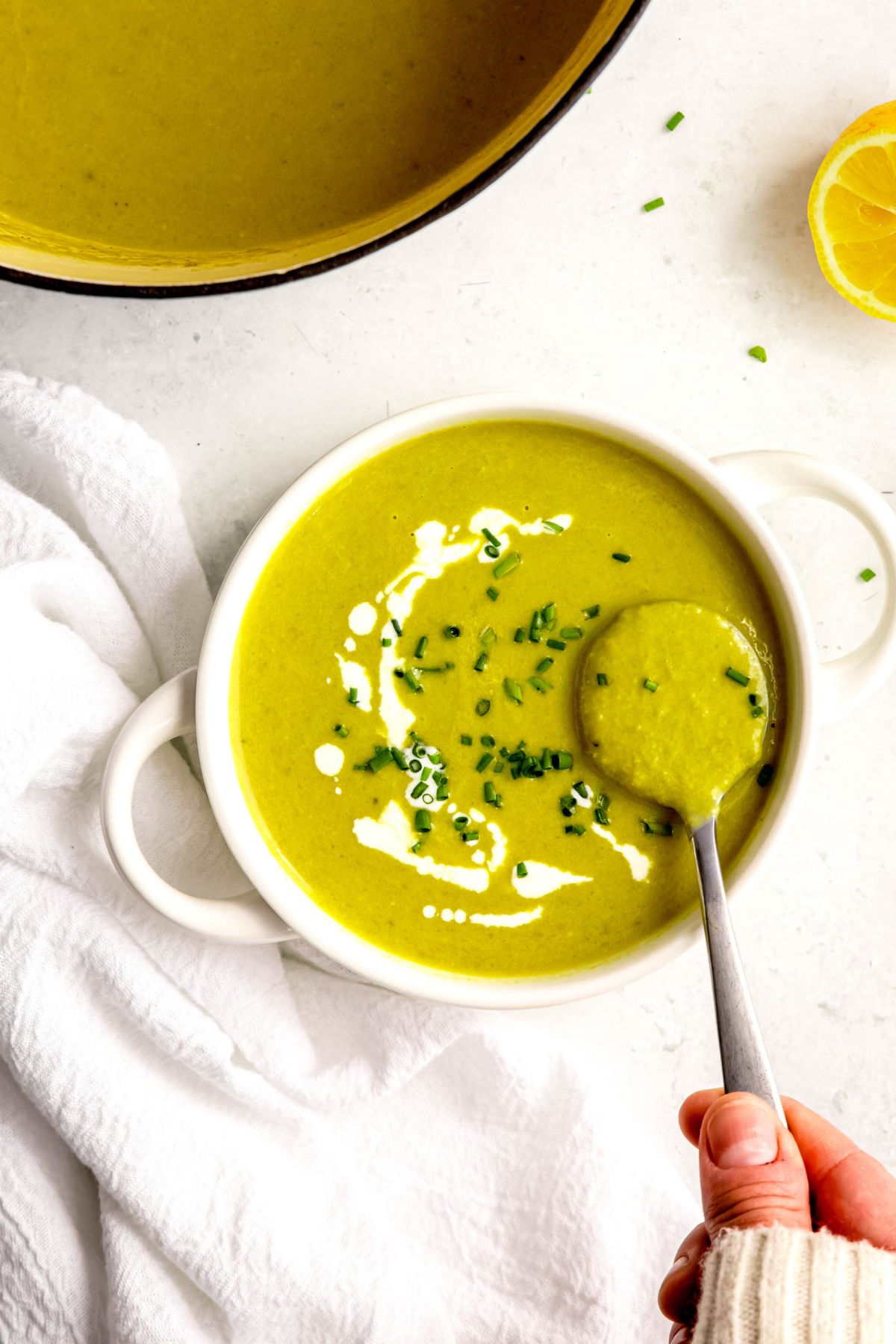 overhead shot of a white soup bowl with handles filled with a serving of creamy asparagus soup garnished with fresh chives and a drizzle of cream; a hand in a white sweater is using a silver spoon to take a bite.