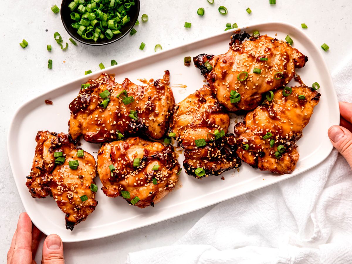 horizontally framed hero shot of hands setting down a rectangular white serving platter with grilled chicken teriyaki thighs on a white table with a white linen and a black pinch bowl overflowing with scallion greens.