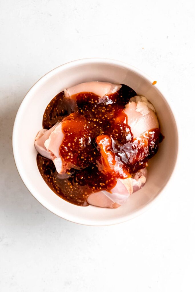 raw boneless skinless chicken thighs in a large white mixing bowl topped with most of the teriyaki sauce as marinade.