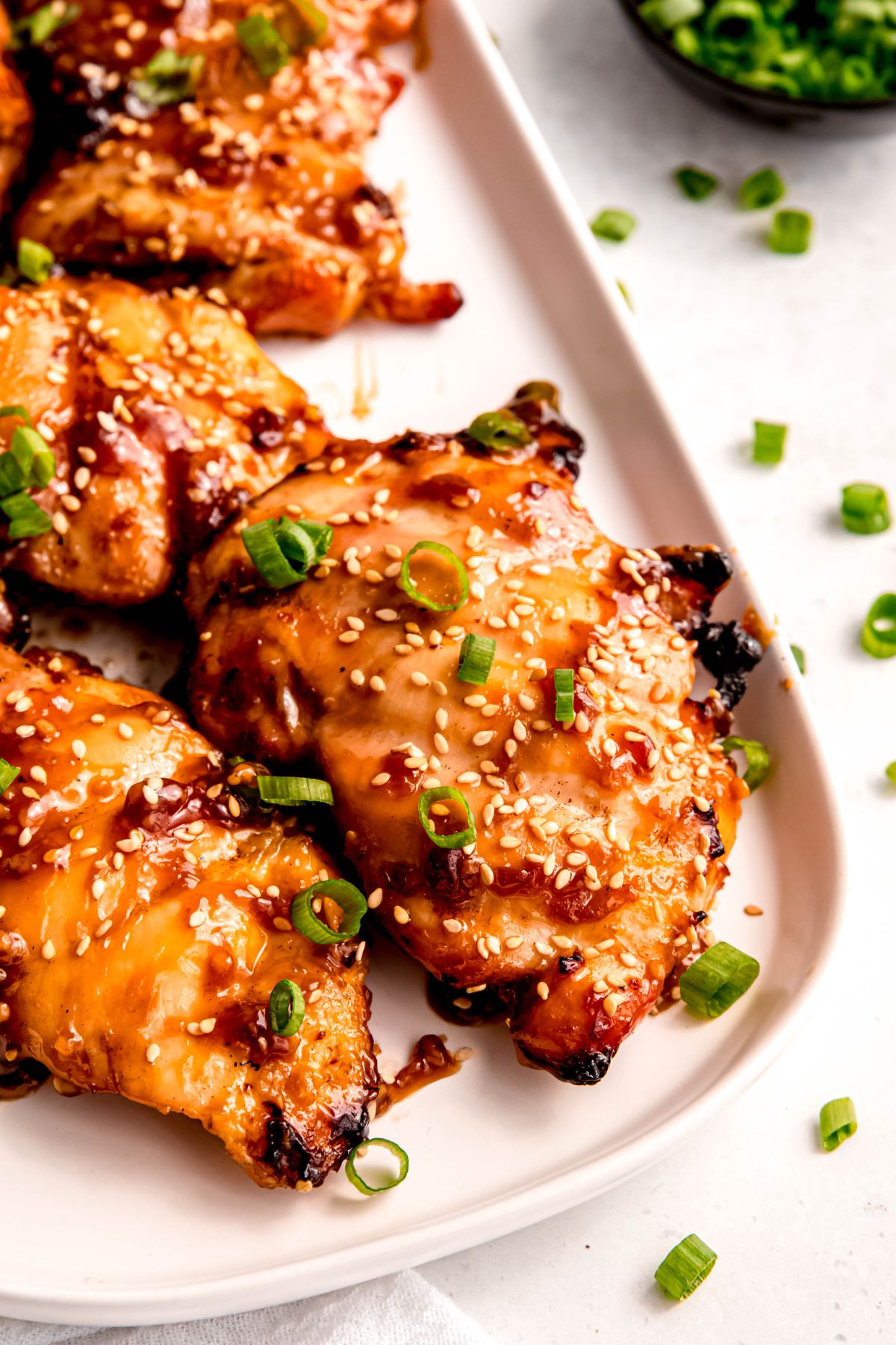 45 degree closeup of the bottom right corner of a rectangular white serving platter with grilled chicken teriyaki thighs topped with green onions and sesame seeds on a white table.