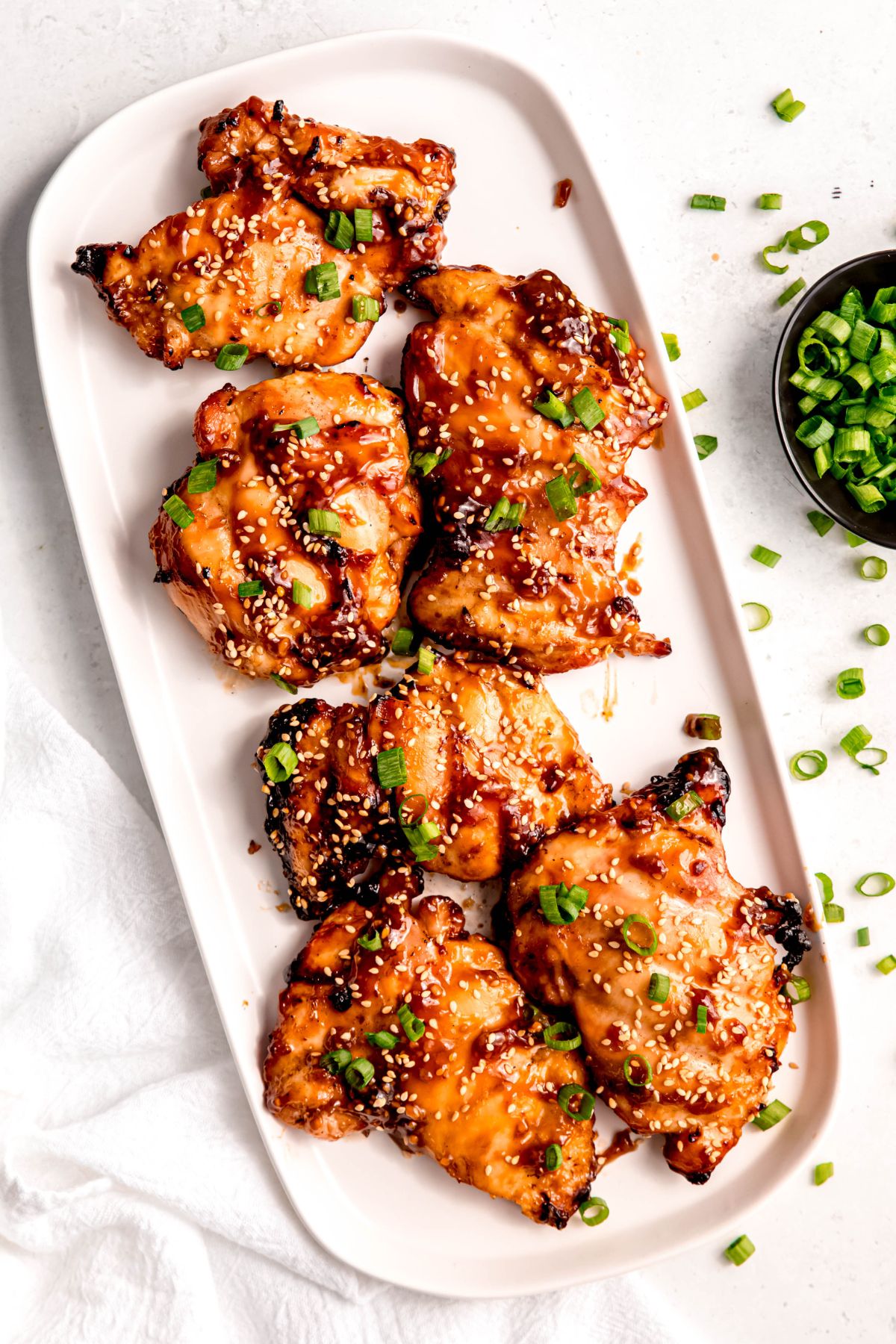 overhead shot of a white oblong serving platter with grilled teriyaki chicken thighs topped with fresh scallions and toasted sesame seeds on a white table.