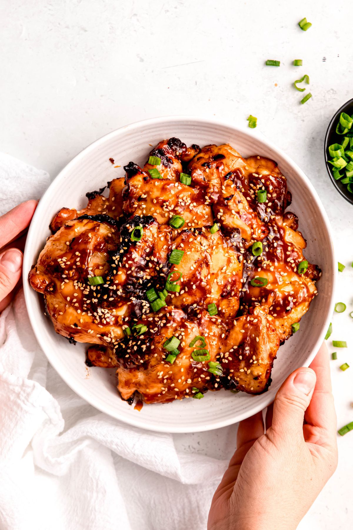 hands setting down a white bowl filled with grilled teriyaki chicken thighs topped with scallion greens and toasted sesame seeds on a white table with a white linen and more scallions scattered around.