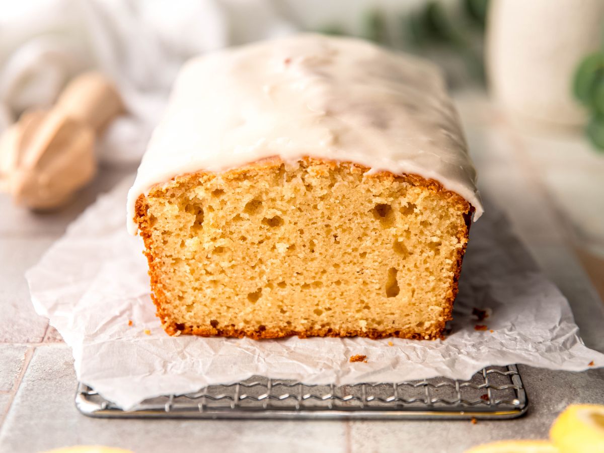 horizontally framed hero shot of a whole copycat Starbucks iced lemon loaf cake on a piece of crumpled white parchment paper on top of a silver wire cooling rack on a grey slate tile table.