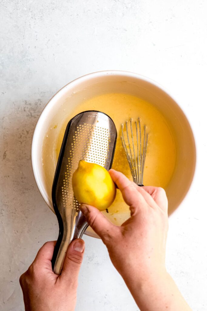 action shot of a hand zesting a lemon over the whisked wet ingredients in the bowl.