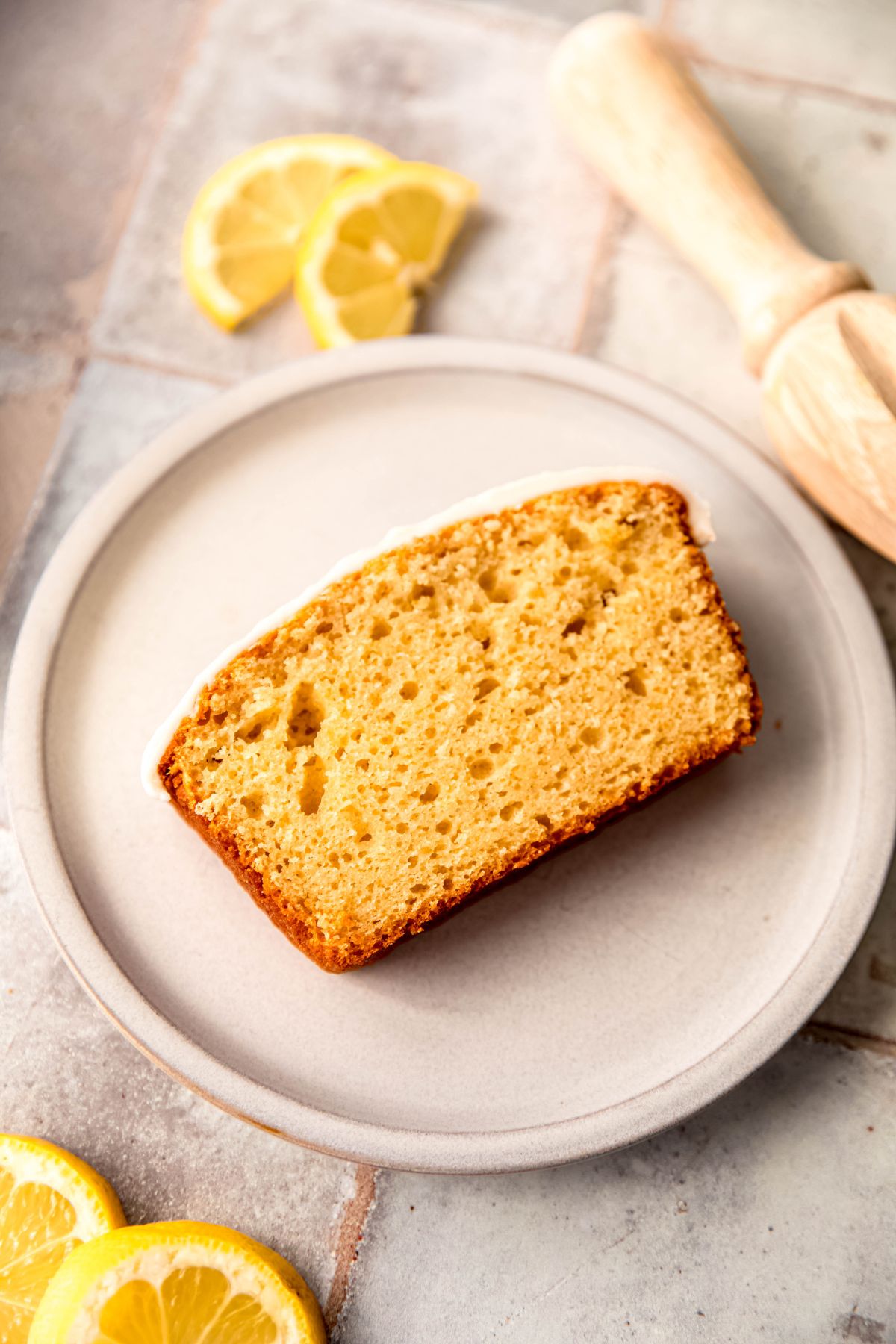 slice of iced lemon loaf cake on a white plate on a slate tile table with slices of fresh lemon and a wooden lemon reamer.