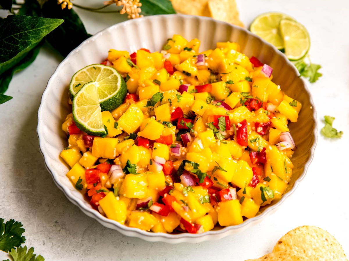 horizontally framed hero shot of colorful mango salsa on a white table with fresh cilantro leaves and yellow corn tortilla chips.