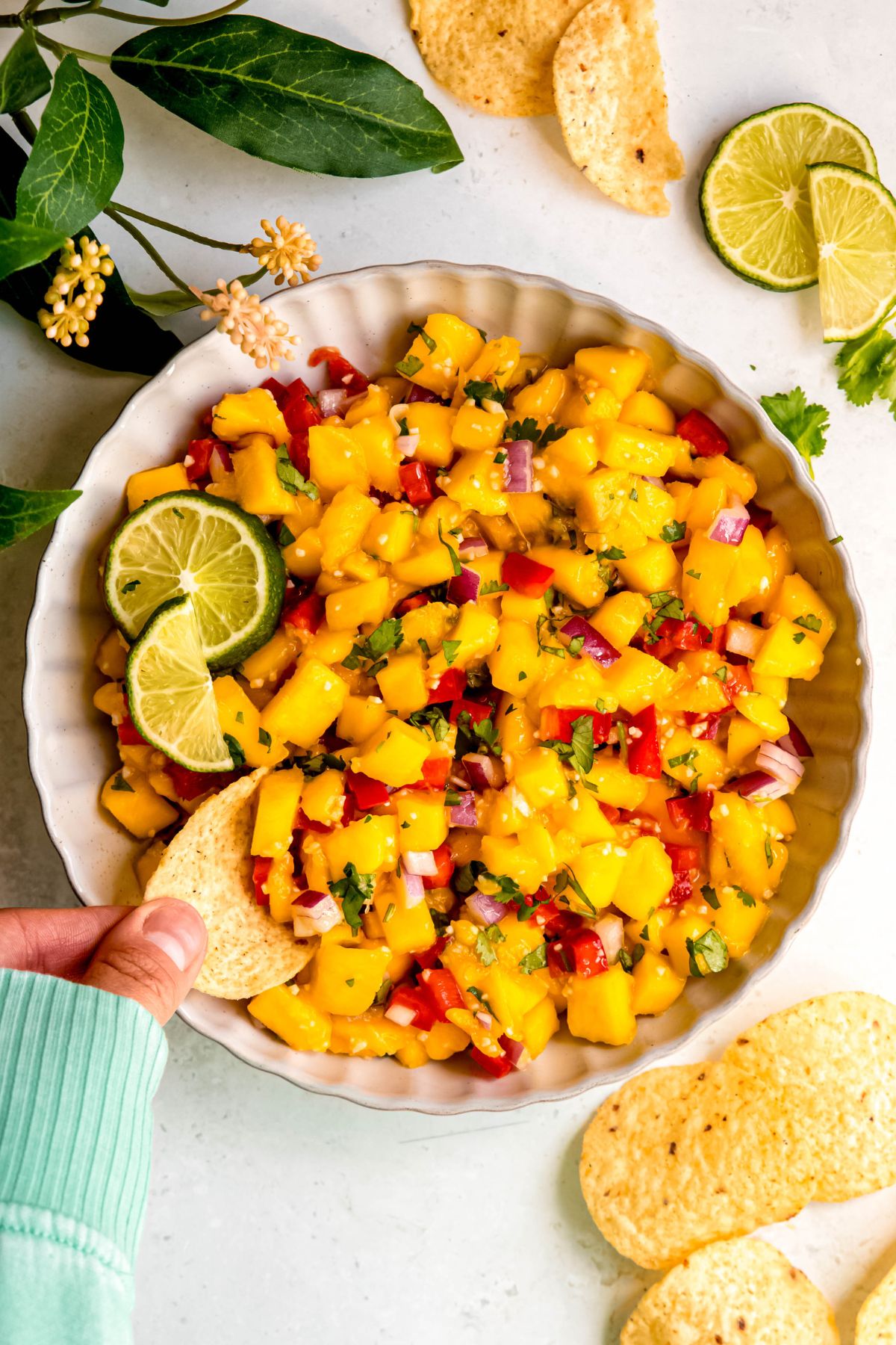 overhead shot of a hand in a mint green sweater up to the knuckles dipping a tortilla chip into a bowl of fresh mango jalapeno salsa on a white table.