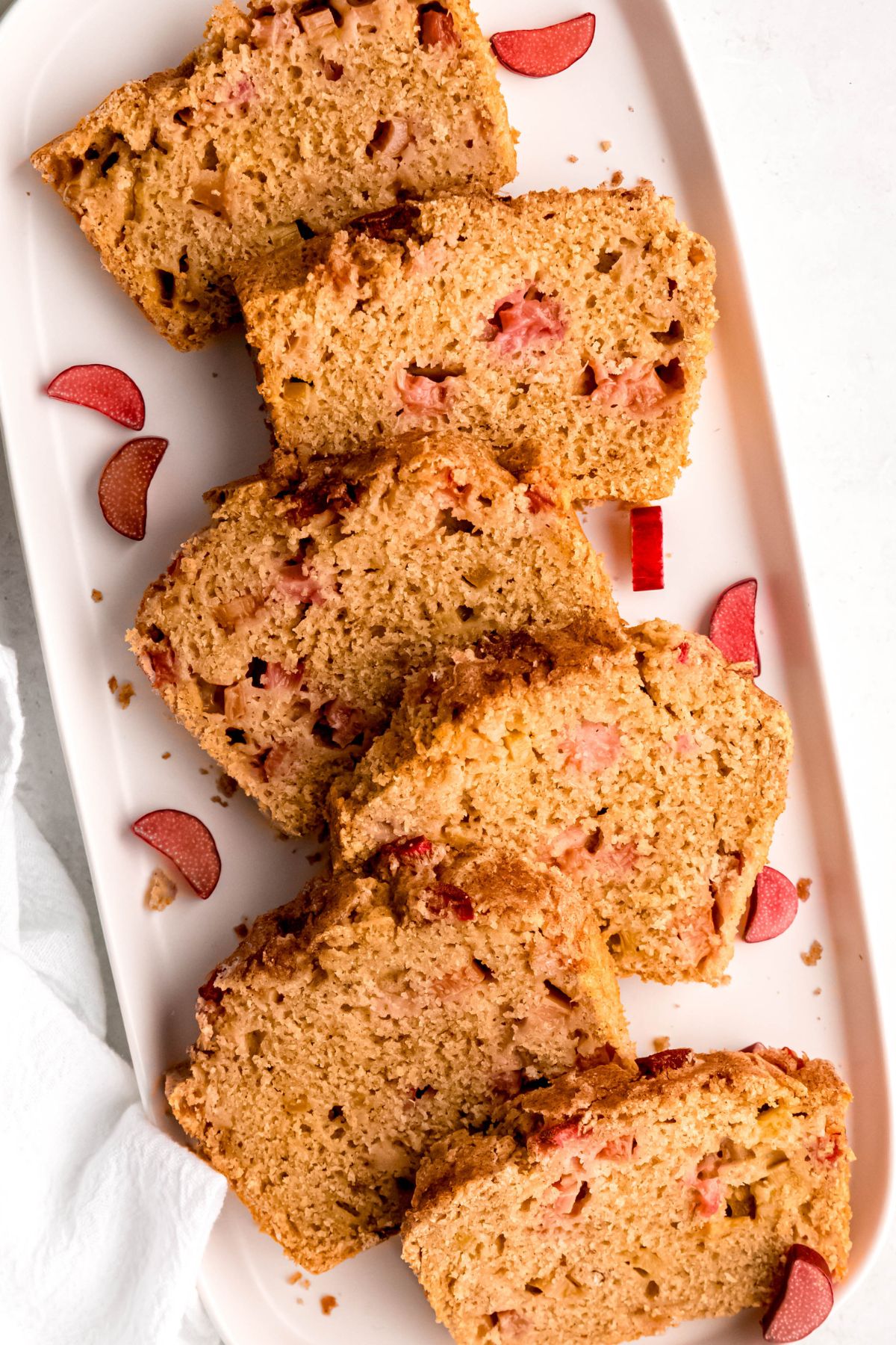 rectangular white serving platter with slices of cinnamon sugar rhubarb bread alternating between the left and right side of the plate, with slices of fresh rhubarb.