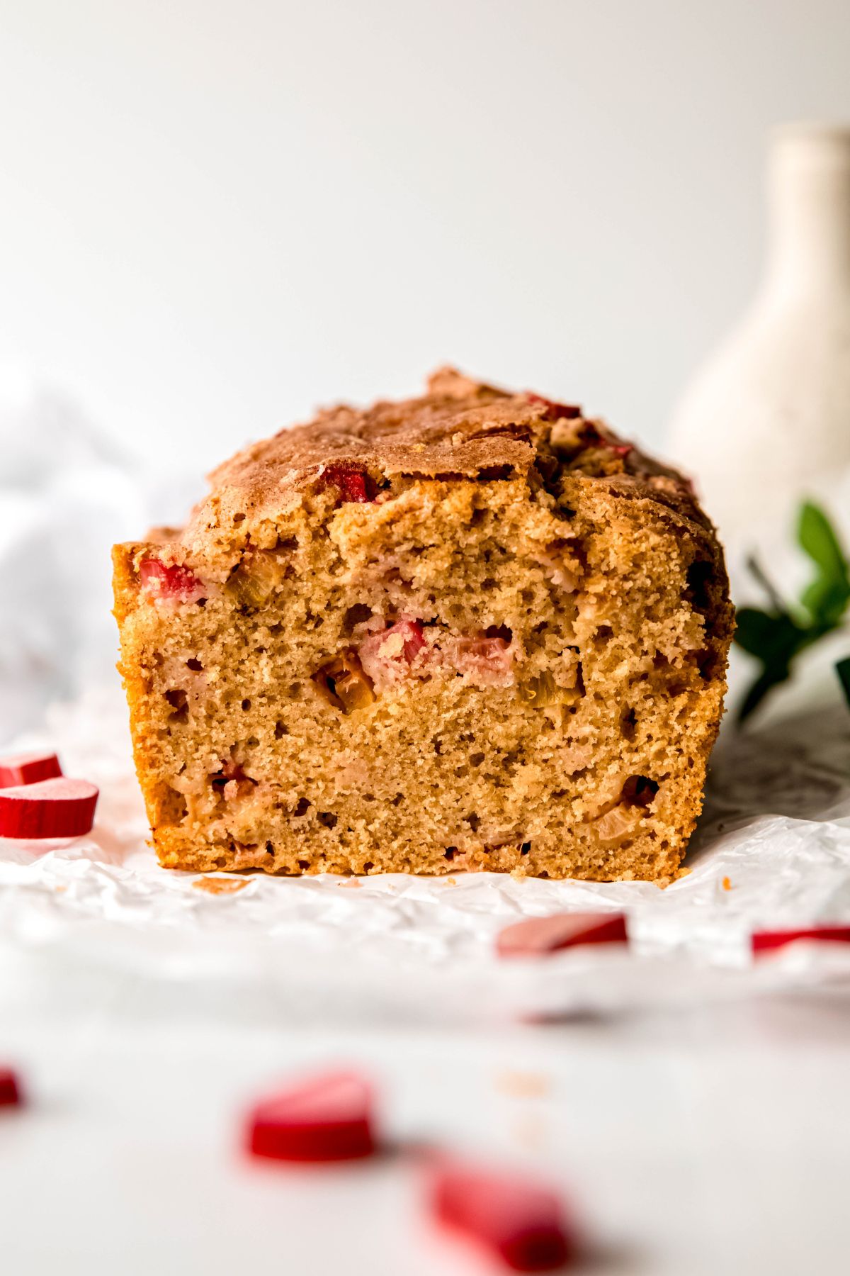 loaf of rhubarb quick bread on a crumpled piece of white parchment paper with fresh pieces of rhubarb scattered around.