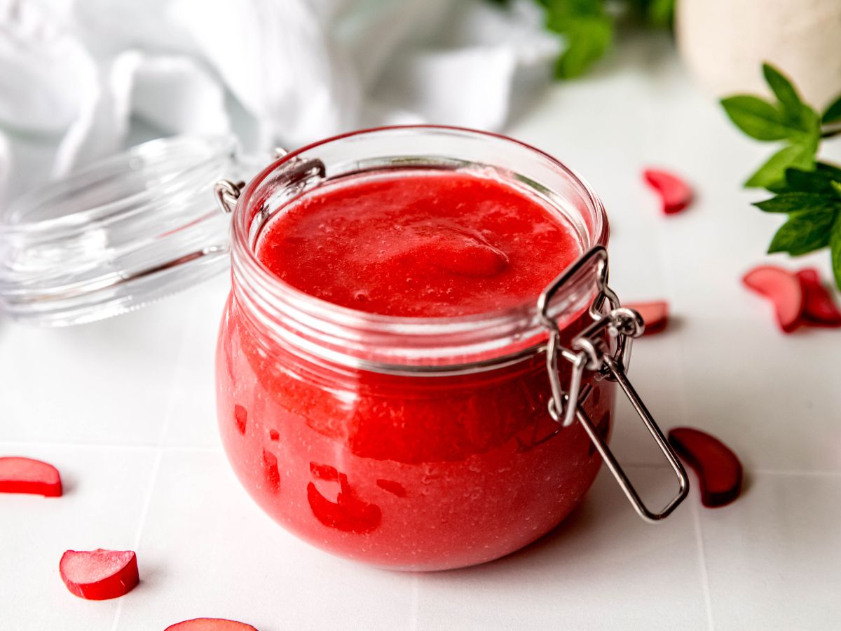 horizontally framed hero image of a jar of homemade orange rhubarb sauce on a white table with a green houseplant, white linen, and red slices of fresh rhubarb.