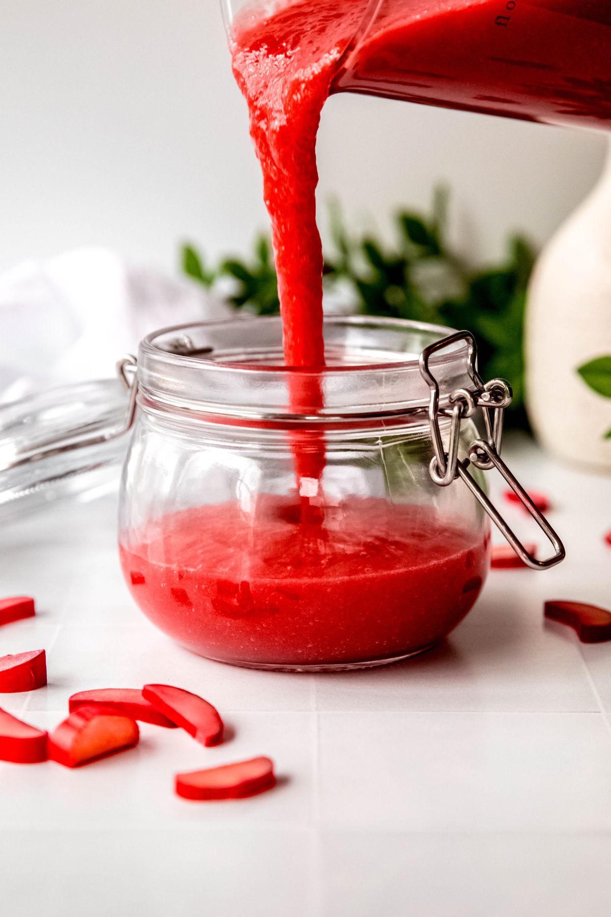 action shot of sweet tart rhubarb sauce being poured into a flip-top mason jar on a white table with a green houseplant blurred out behind and fresh slices of rhubarb in the fore.