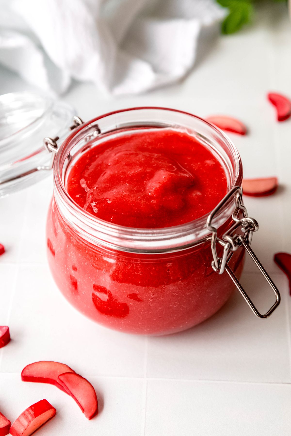 open flip top jar of bright red blended rhubarb sauce on a white table with slices of fresh rhubarb scattered around.