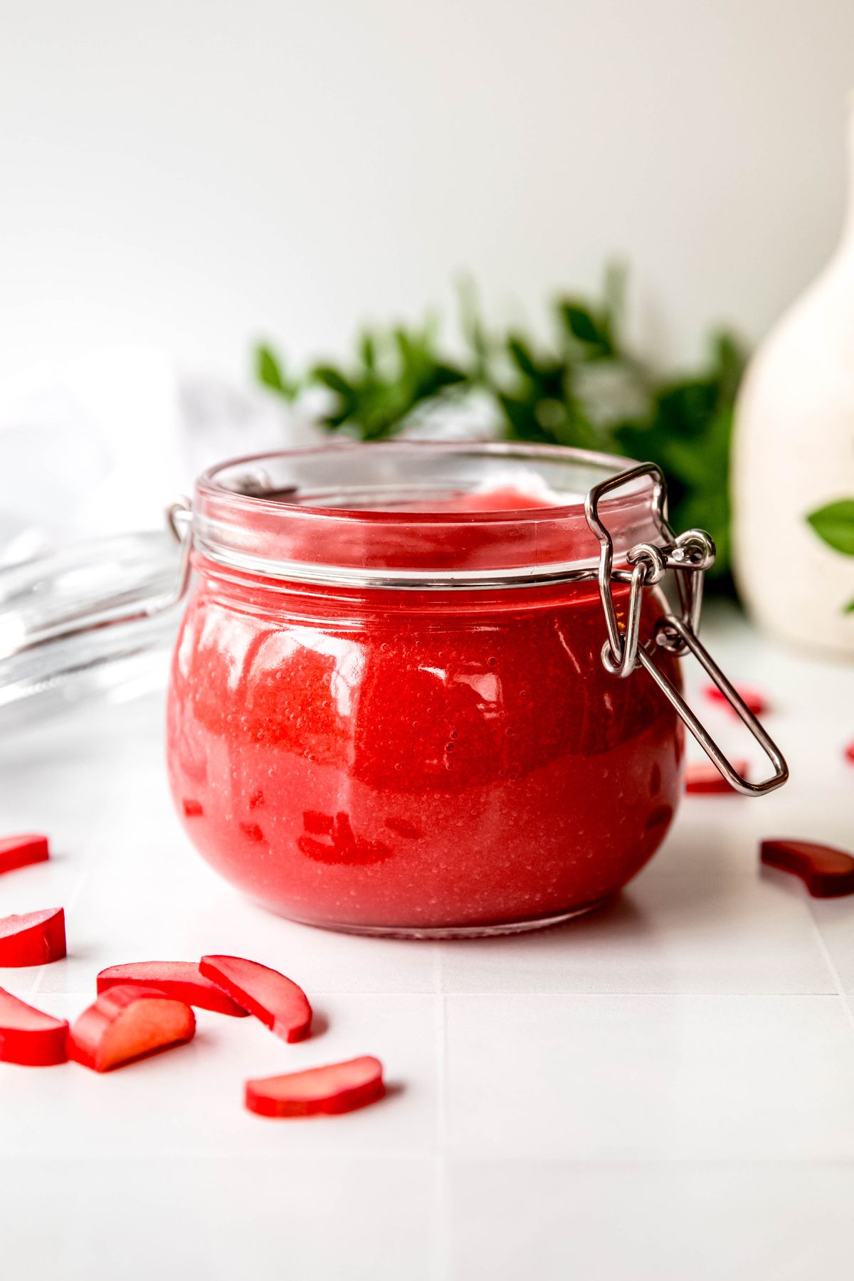 side on view of a clear french style mason jar filled with sweet rhubarb sauce on a white table with a green houseplant and white jug blurred out in the background.