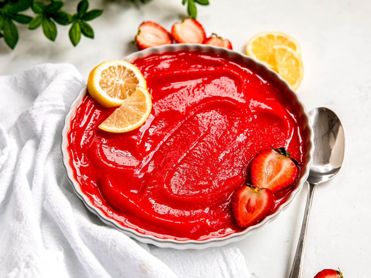 horizontally framed 45 degree hero shot of a bowl of homemade strawberry sorbet topped with a halved strawberry and two lemon slices on a white table with a white linen, a silver spoon, and more sliced fruit.