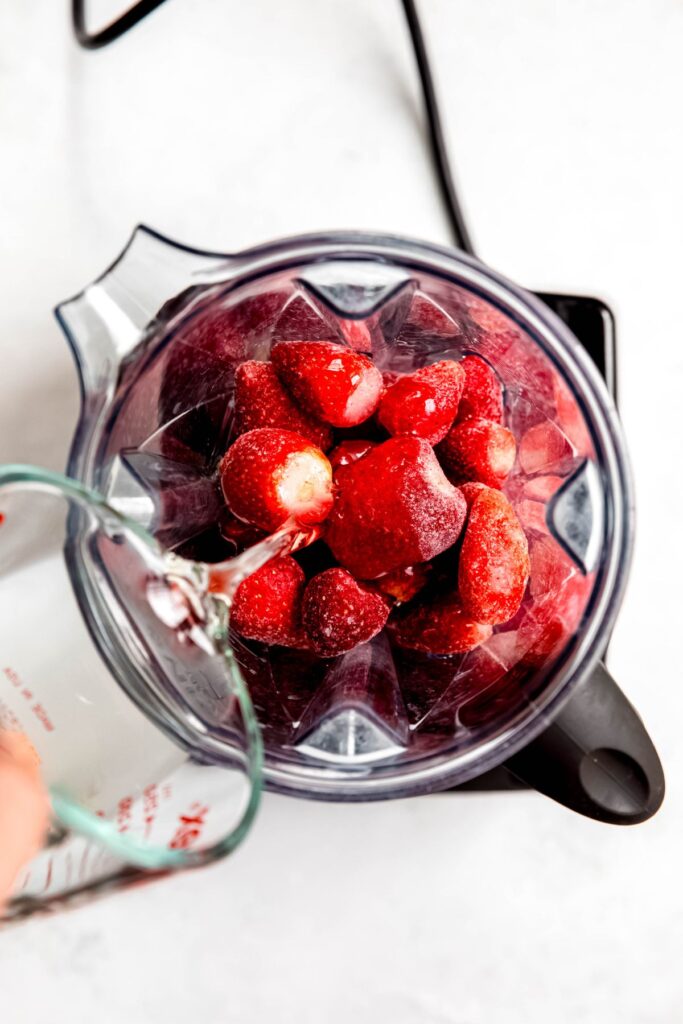 action shot of warm water being poured into a blender with frozen strawberries and simple syrup.