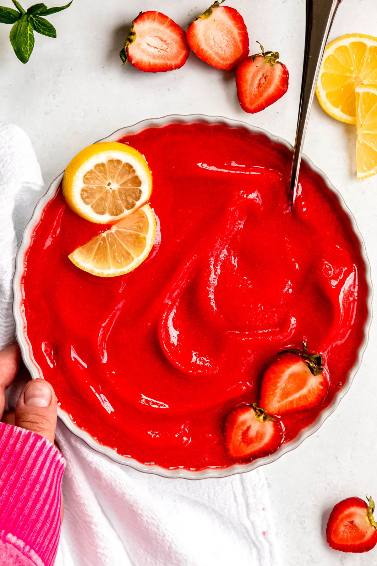 hand in a pink sweater up to the knuckles setting down a white bowl of bright red strawberry sorbet on a white table with sliced berries.